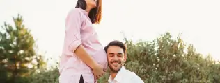 Homem sorridente apoiando a cabeça na barriga de grávida da parceira em um parque.