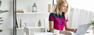 Mulher loira sorrindo enquanto trabalha no laptop em uma mesa com uma caneca.