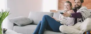 Casal sorrindo e assistindo TV junto no sofá, cobertos por uma manta.